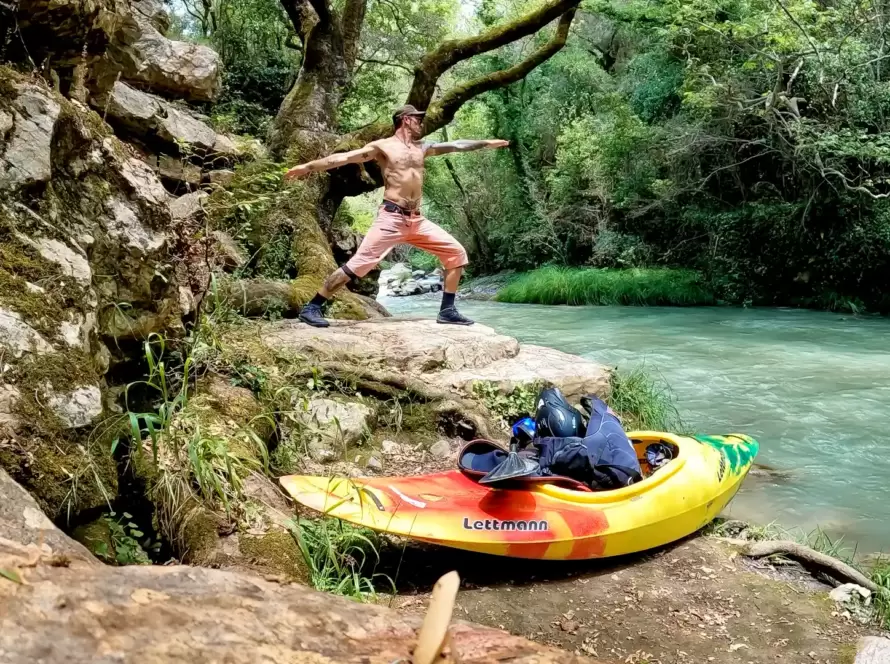 A man practices yoga next to a river in Greece with a kayak in the foreground