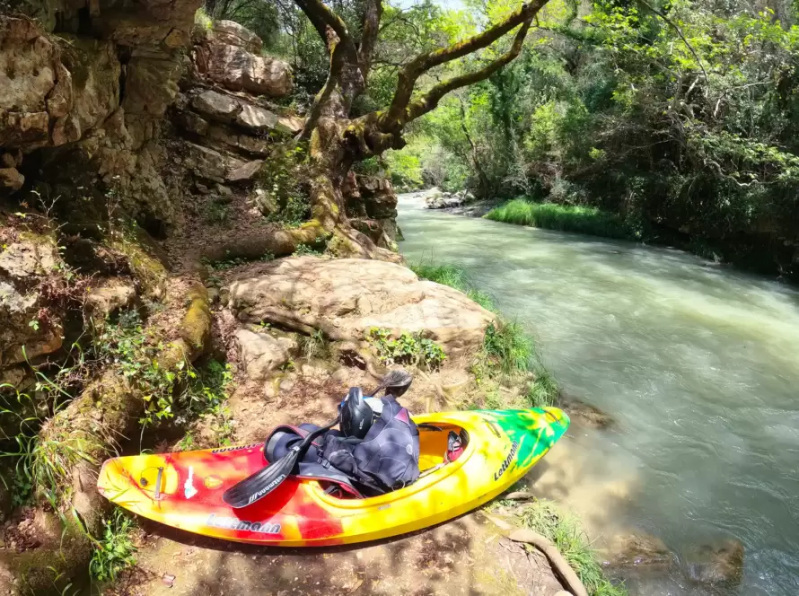 A kayak lays on the ground next to a river in Greece.