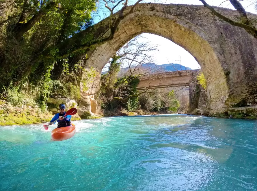 A person kayaks in a river, with an ancient Greek bridge in the background.