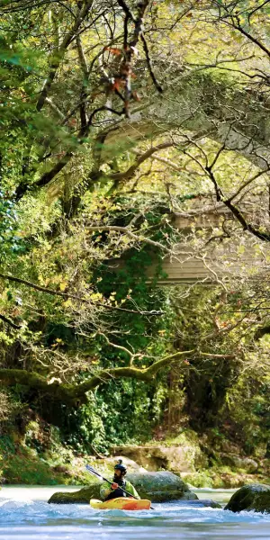 A person kayaks in a river under an ancient Greek bridge