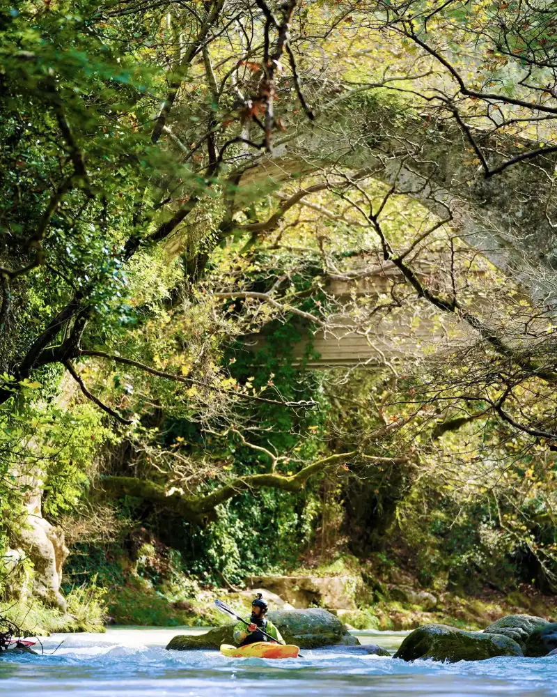 A person kayaks in a river under an ancient Greek bridge